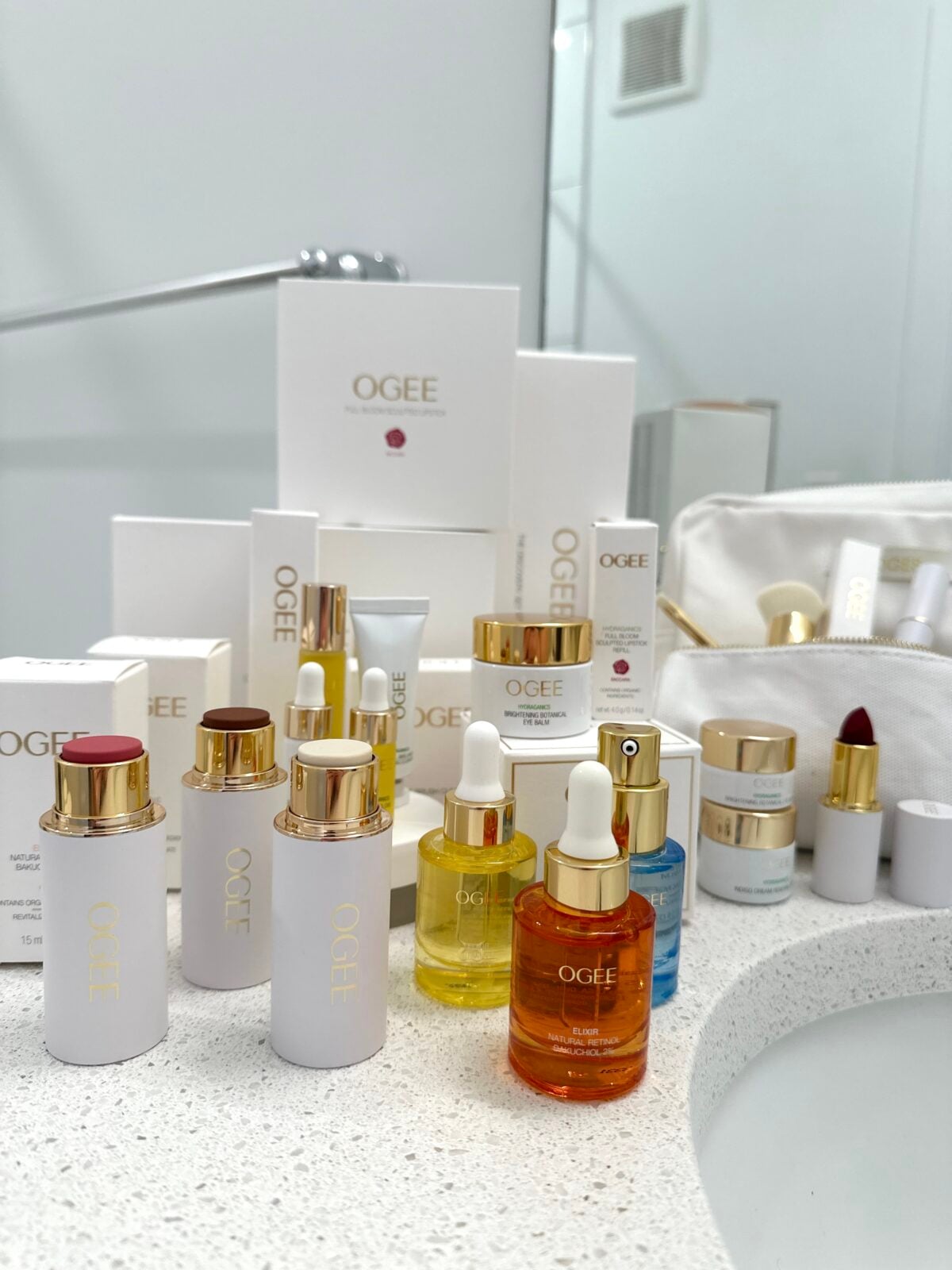 A collection of OGEE skincare and beauty products, including serums, creams, lipsticks, and dropper bottles, arranged on a white bathroom countertop near a sink and a white cosmetic bag.