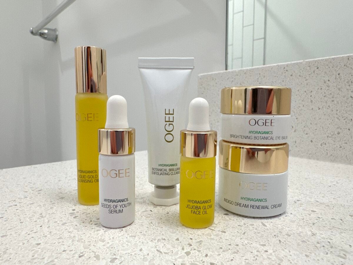 Six skincare products from OGEE are arranged on a speckled bathroom counter. Bottles, jars, and a tube in white and gold packaging, labeled with product names like cleanser, exfoliating gel, face oil, and renewal cream.