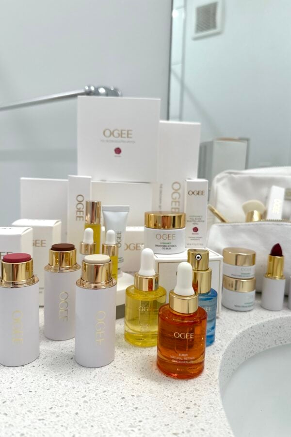 A collection of OGEE skincare and beauty products, including serums, creams, lipsticks, and dropper bottles, arranged on a white bathroom countertop near a sink and a white cosmetic bag.