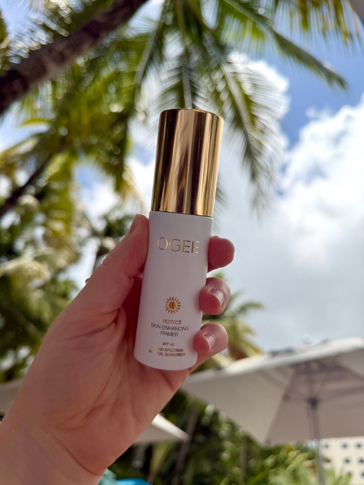 A hand holds a white and gold bottle of OGEE Tinted Skin Enhancing Primer SPF 30 outdoors, with palm trees, a blue sky, and a sun umbrella in the background.