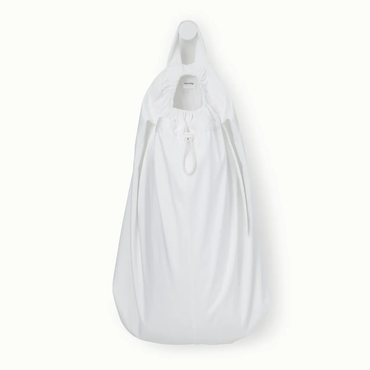 A white cotton laundry bag with a drawstring closure is hanging on a white wall hook. The bag has a rounded bottom and a small opening at the top, with a visible brand tag inside the rim.