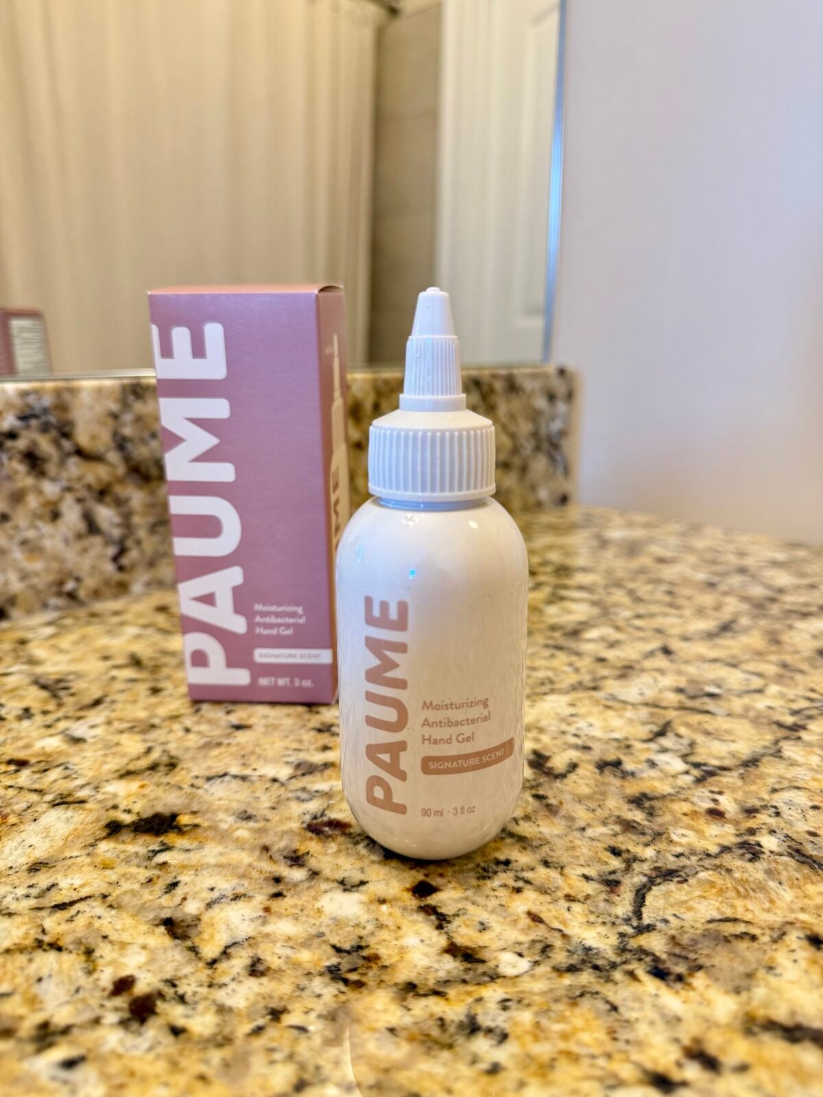 A bottle of PAUME Moisturizing Antibacterial Hand Gel sits on a granite bathroom counter with its pink box standing upright in the background near a mirror.