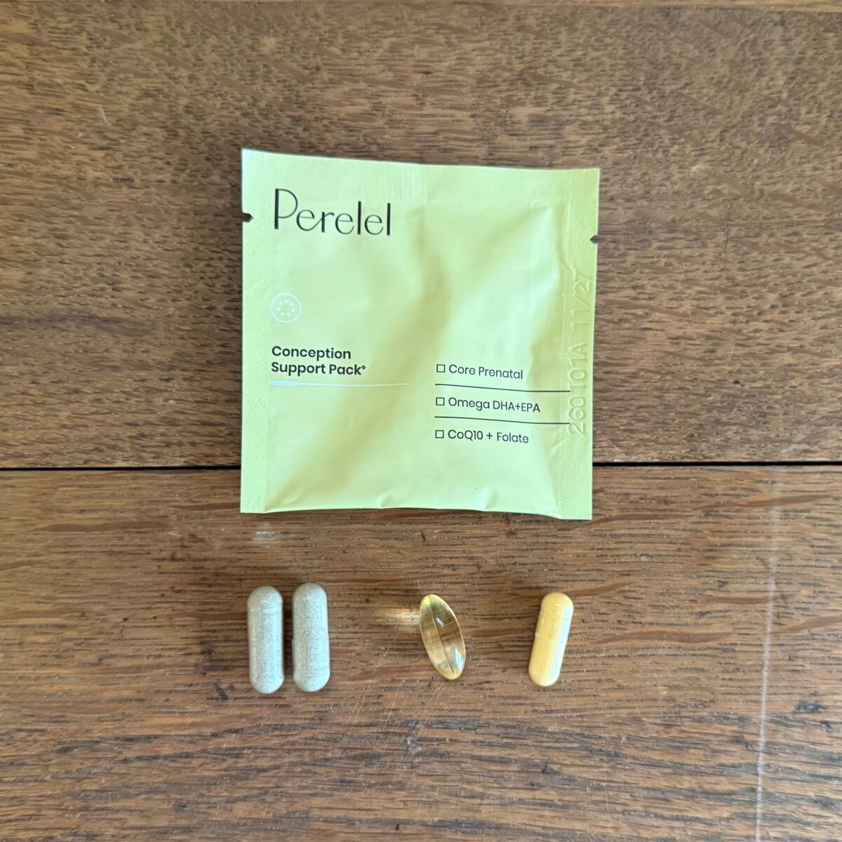 A yellow Perlele Conception Support Pack lies on a wooden surface. Below it are three capsules—two gray, one yellow—and one clear, oval softgel.