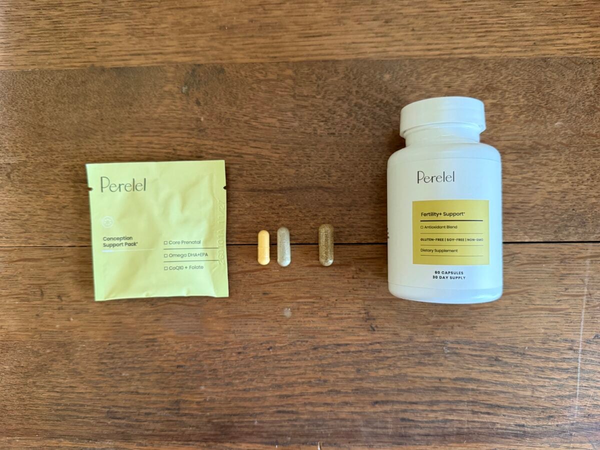 A yellow Perel packet, three assorted capsules, and a white Perel Fertility Support bottle with a yellow label are arranged on a wooden surface.