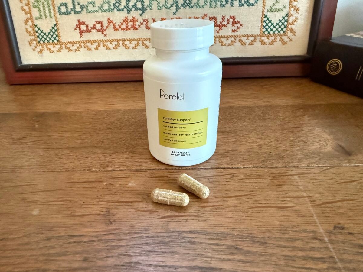 A white bottle labeled “Perelel Femten+ Support” sits on a wooden surface with three beige capsules in front. There is a framed embroidered artwork and a book in the background.