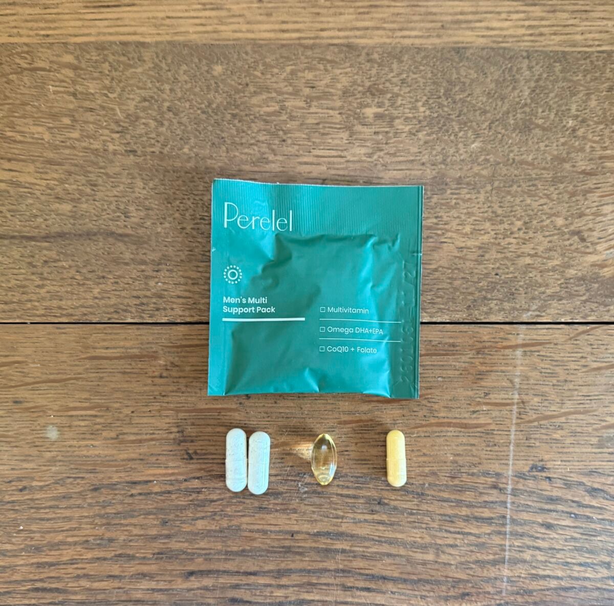 A green packet labeled Perelel Mens Multi Support Pack is on a wooden surface. Below it are four pills: two white oval-shaped, one large transparent oval, and one yellow capsule.