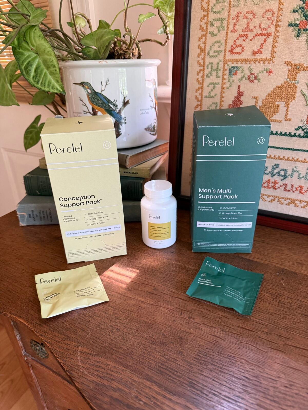 Two Perel products, Conception Support Pack and Man’s Multi Support Pack, are displayed on a wooden table with their boxes, packets, and a white bottle. Decorative plants and framed embroidery are in the background.