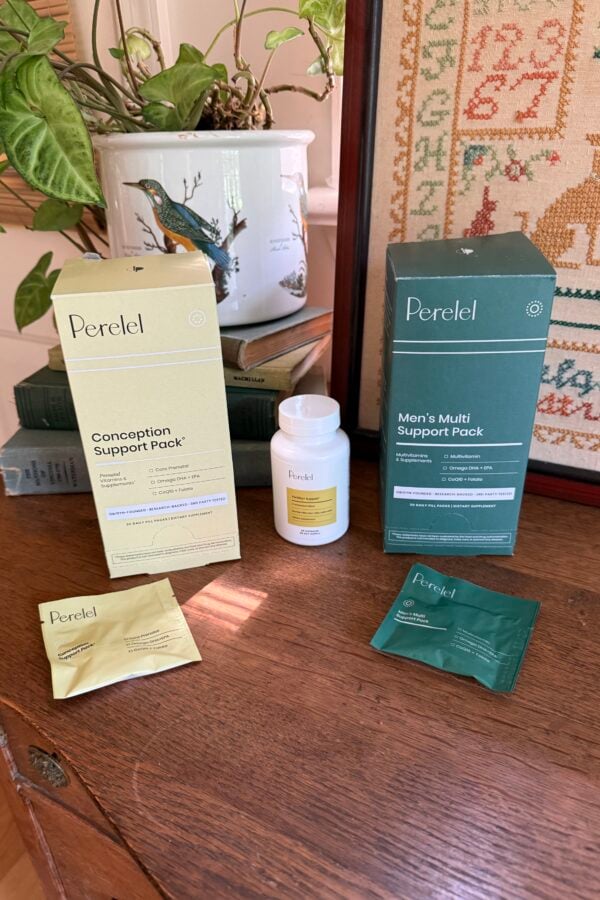 Two Perel products, Conception Support Pack and Man’s Multi Support Pack, are displayed on a wooden table with their boxes, packets, and a white bottle. Decorative plants and framed embroidery are in the background.