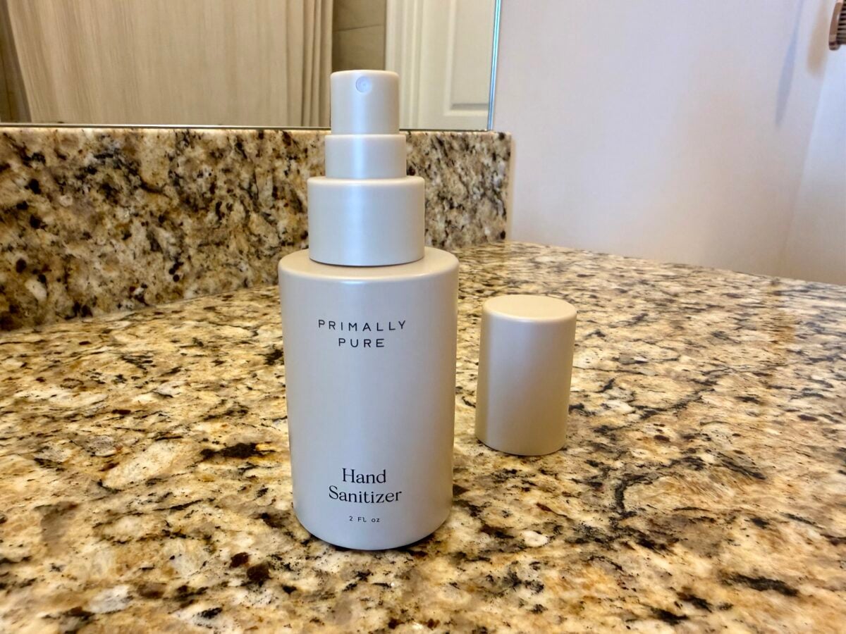 A beige bottle labeled Primally Pure Hand Sanitizer sits on a granite countertop with its cap off. A mirror and door are visible in the background.