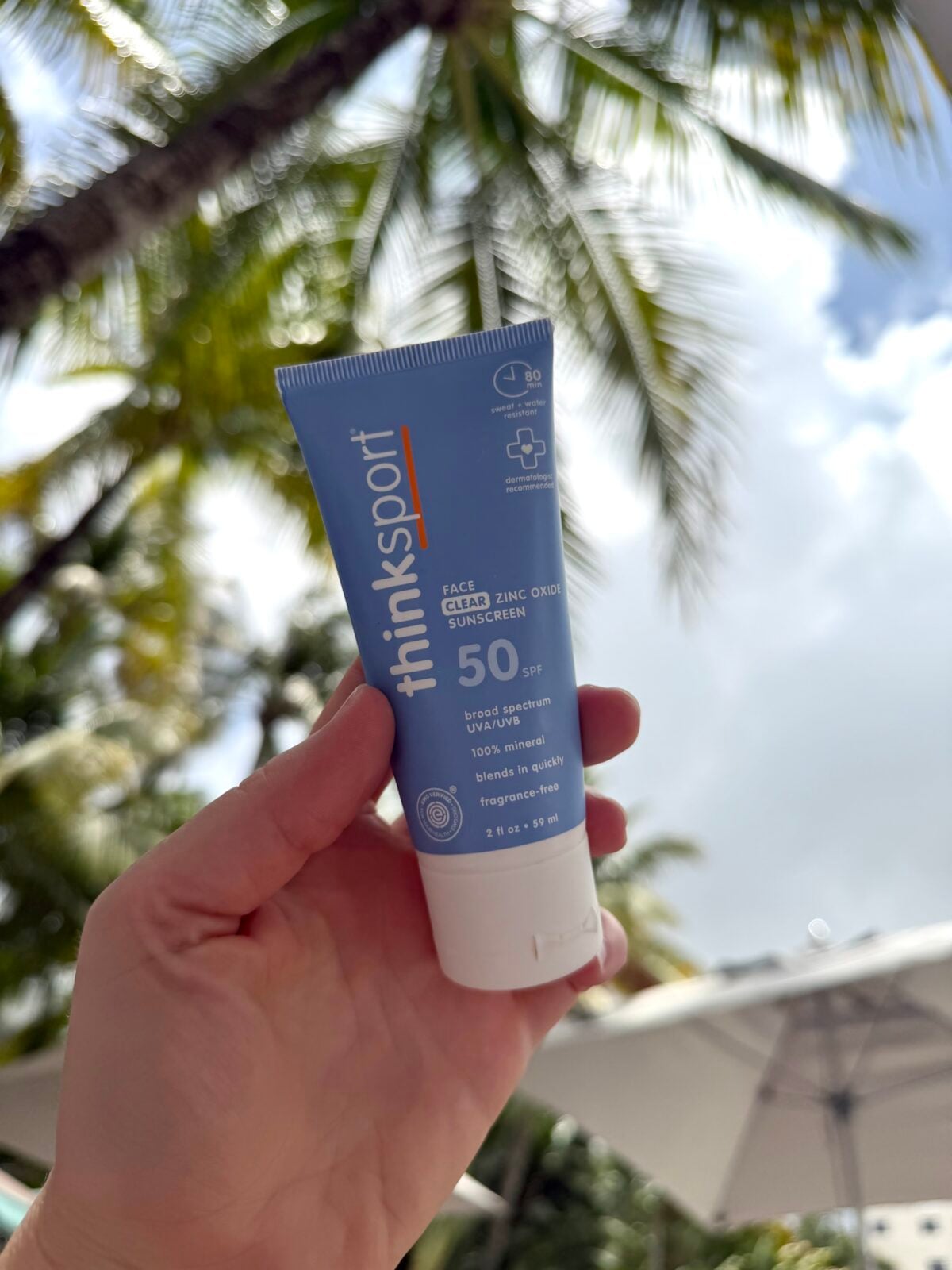 A hand holds a tube of Thinksport SPF 50 sunscreen with a background of palm trees and a bright, partly cloudy sky.