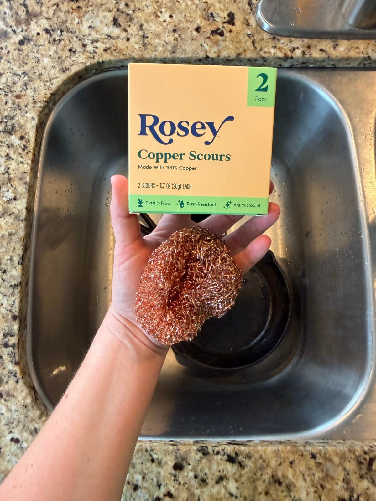A hand holds a copper scouring pad and a box labeled “Rosey Copper Scours” over a kitchen sink. The countertop is granite, and the package states it contains a 2 pack of plastic-free, antimicrobial pads.