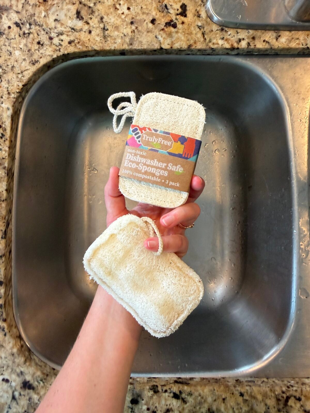 A hand holds two rectangular eco-sponges, one with a brown label reading Dishwasher Safe Eco-Sponges, above a stainless steel sink and a granite countertop.