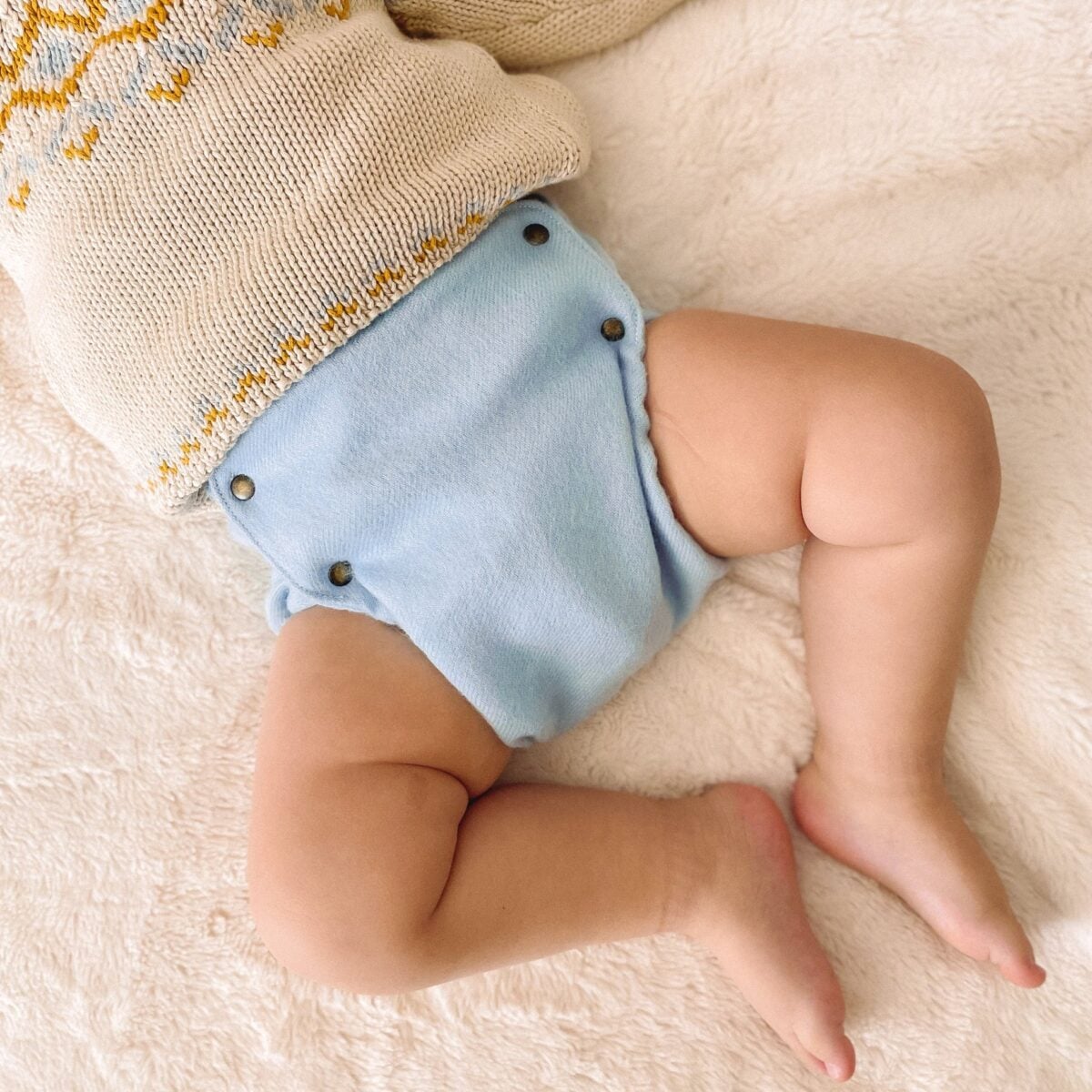 A baby wearing a cream-colored sweater with a yellow pattern and a light blue cloth diaper lies on a soft, beige blanket. The image shows the babys legs and lower torso.