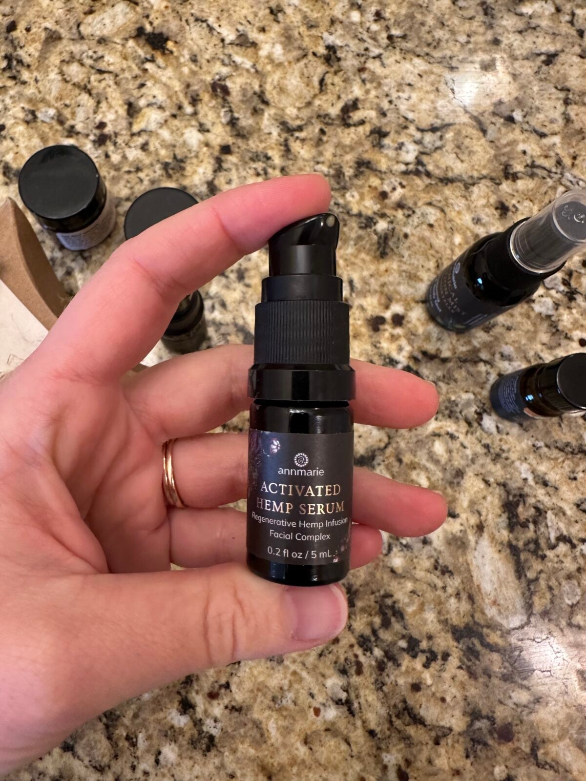 A hand holds a small black bottle labeled Annmarie Activated Hemp Serum against a granite countertop. Other similar bottles are blurred in the background. The bottle contains 0.21 fl oz or 5 mL.