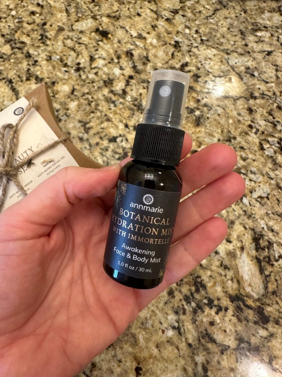 A hand holds a small black bottle labeled Annmarie Botanical Hydration Mist with Immortelle for face and body, against a granite countertop background.