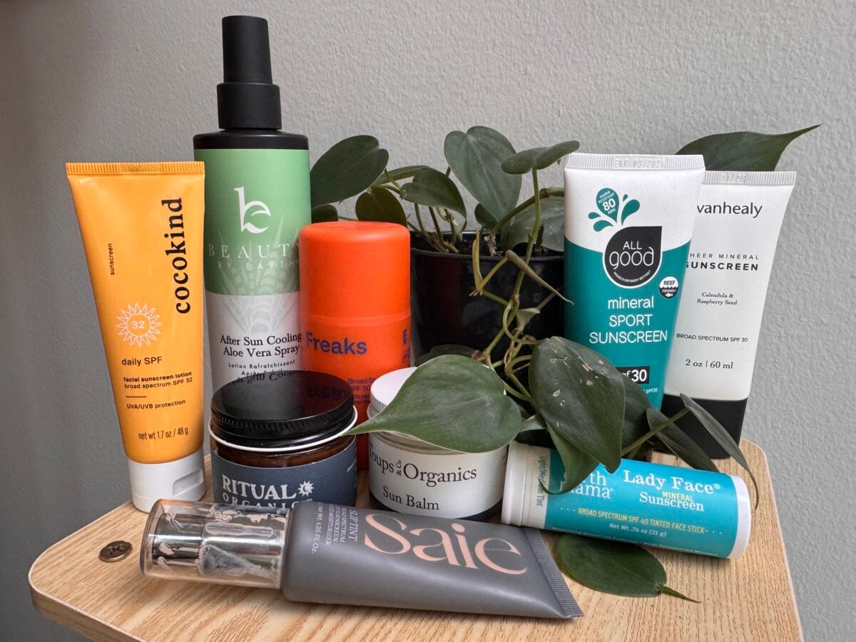 A variety of sunscreen and after-sun care products in different tubes, bottles, and jars are arranged on a wooden table next to a leafy green plant. The background is a plain, light-colored wall.