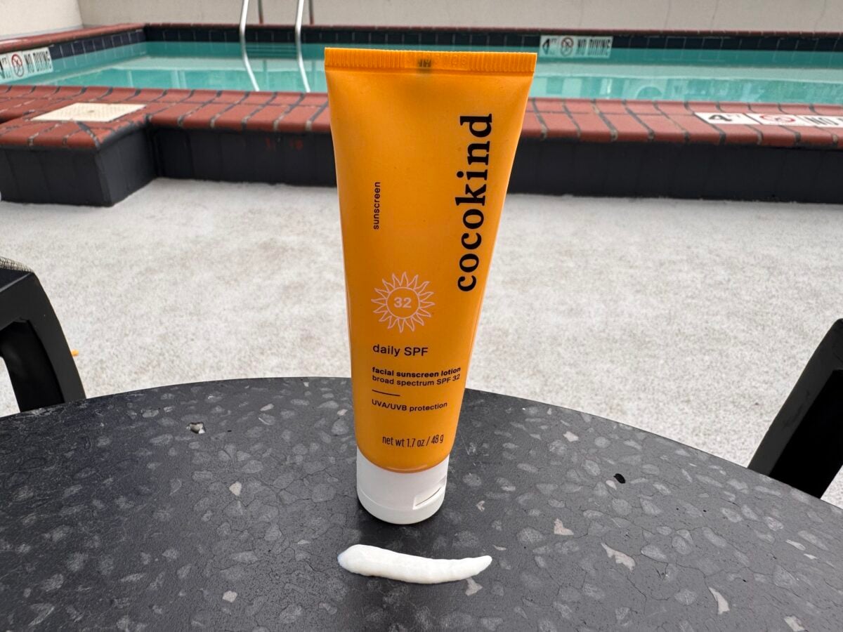 A tube of Cocokind daily SPF 32 sunscreen stands on a round black table with sunscreen squeezed in front of it. A pool and tiled edge are visible in the background.