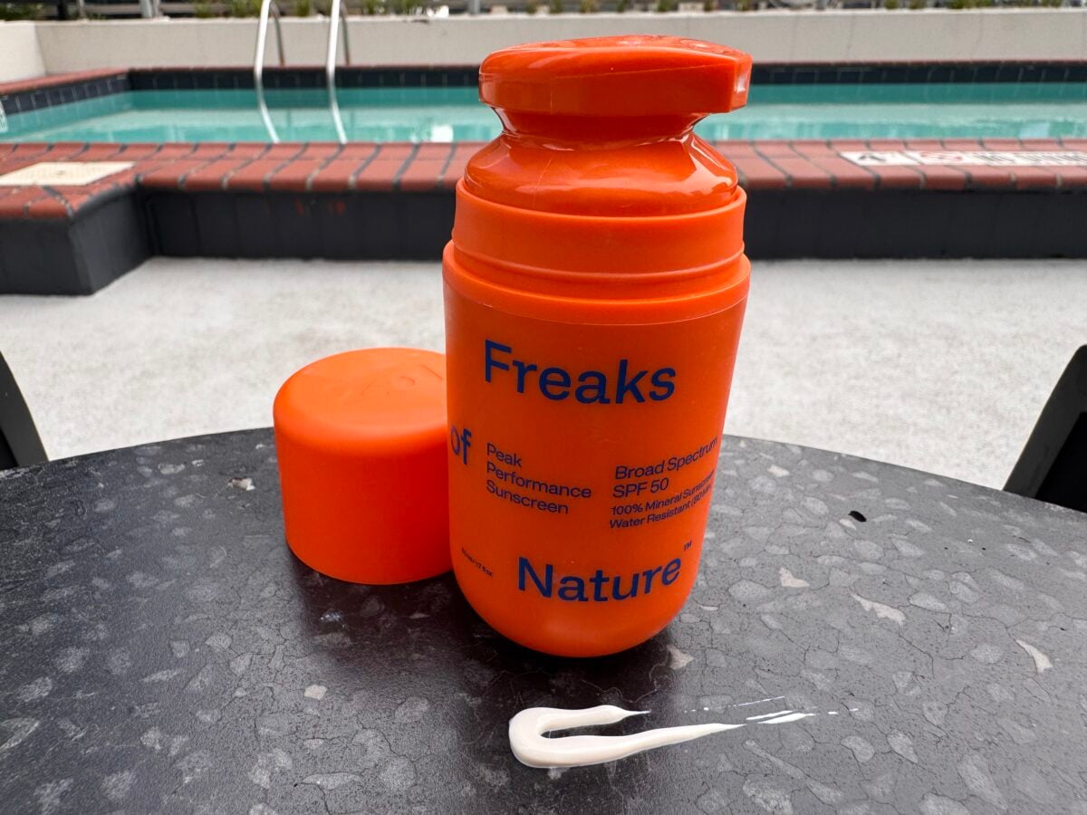 An orange sunscreen bottle labeled Freaks of Nature sits on a black table by a pool. The cap is off, lying nearby, and a line of white sunscreen lotion is spread on the table in front of the bottle.