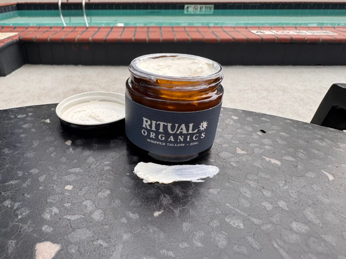 An open jar labeled Ritual Organics Whipped Tallow + Zinc sits on a dark table. The lid is off to the side, with some cream spread on the table. In the background, there is a pool and brick edging.