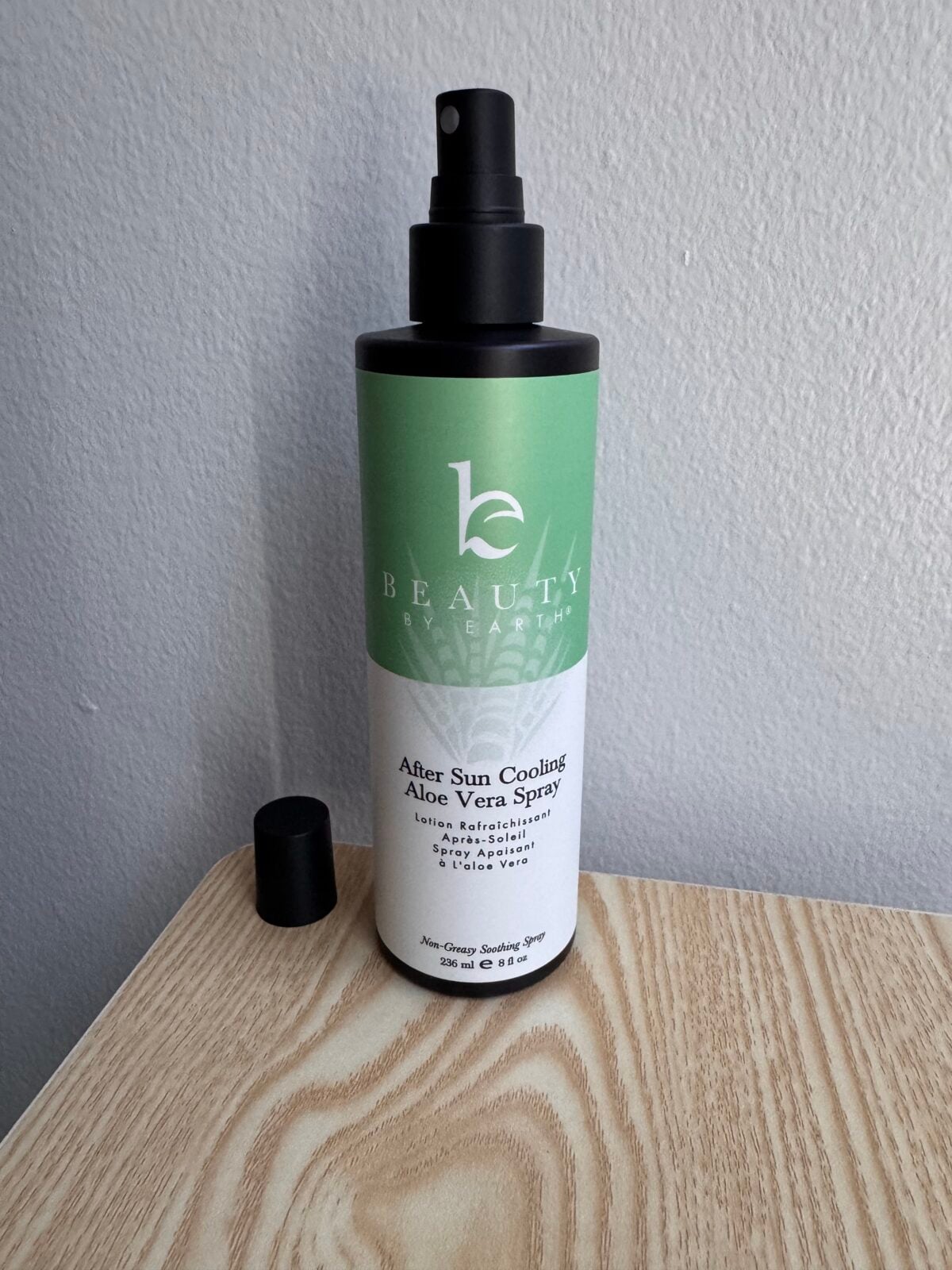A bottle of Beauty Earth After Sun Cooling Aloe Vera Spray sits on a wooden surface near a light-colored wall. The spray cap is attached, and the black cap is placed next to the bottle.