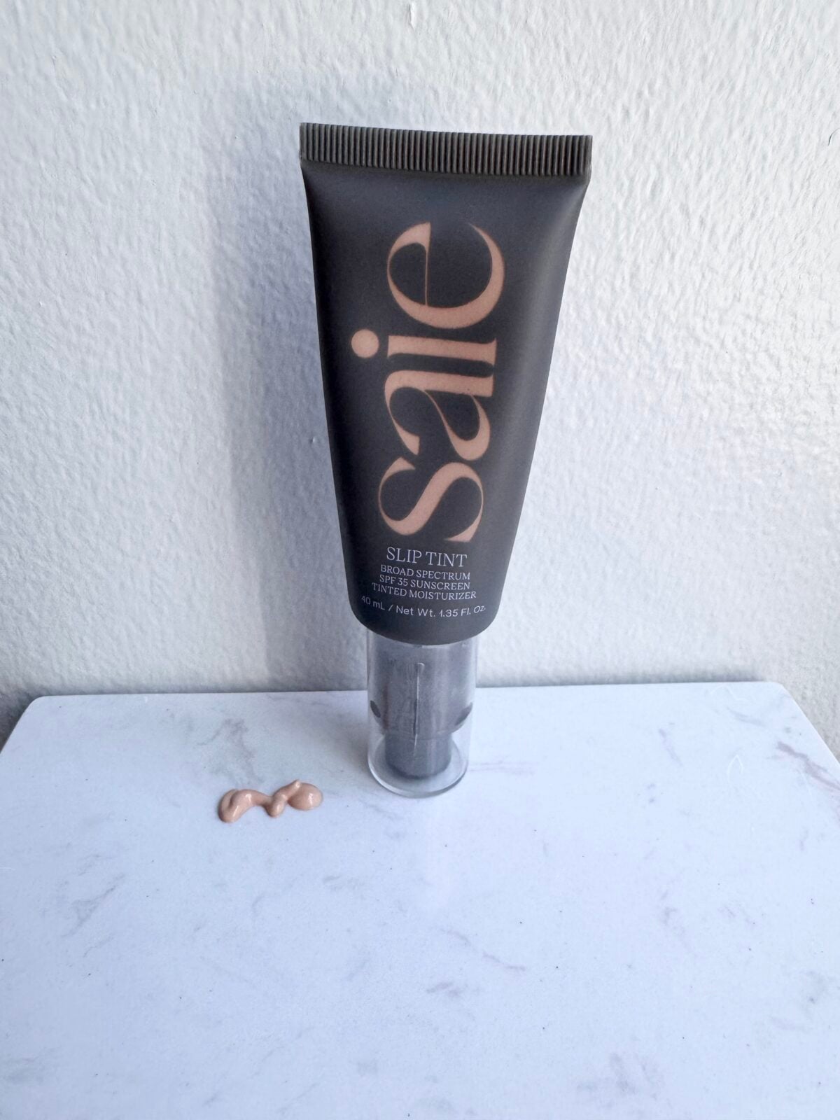 A black tube labeled Saie Slip Tint stands upright on a white surface. Two small blobs of light beige tinted moisturizer are beside it. The background is a plain, light-colored wall.