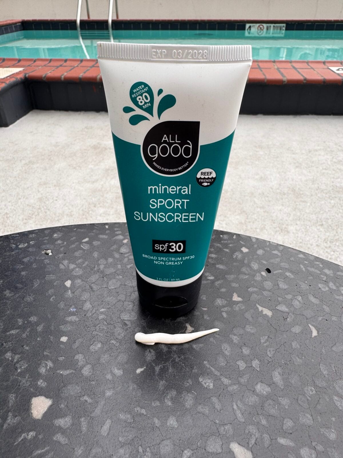 A tube of All Good mineral sport sunscreen SPF 30 stands upright on a round black outdoor table with a seashell in front of it. A pool and part of a pool ladder are visible in the background.