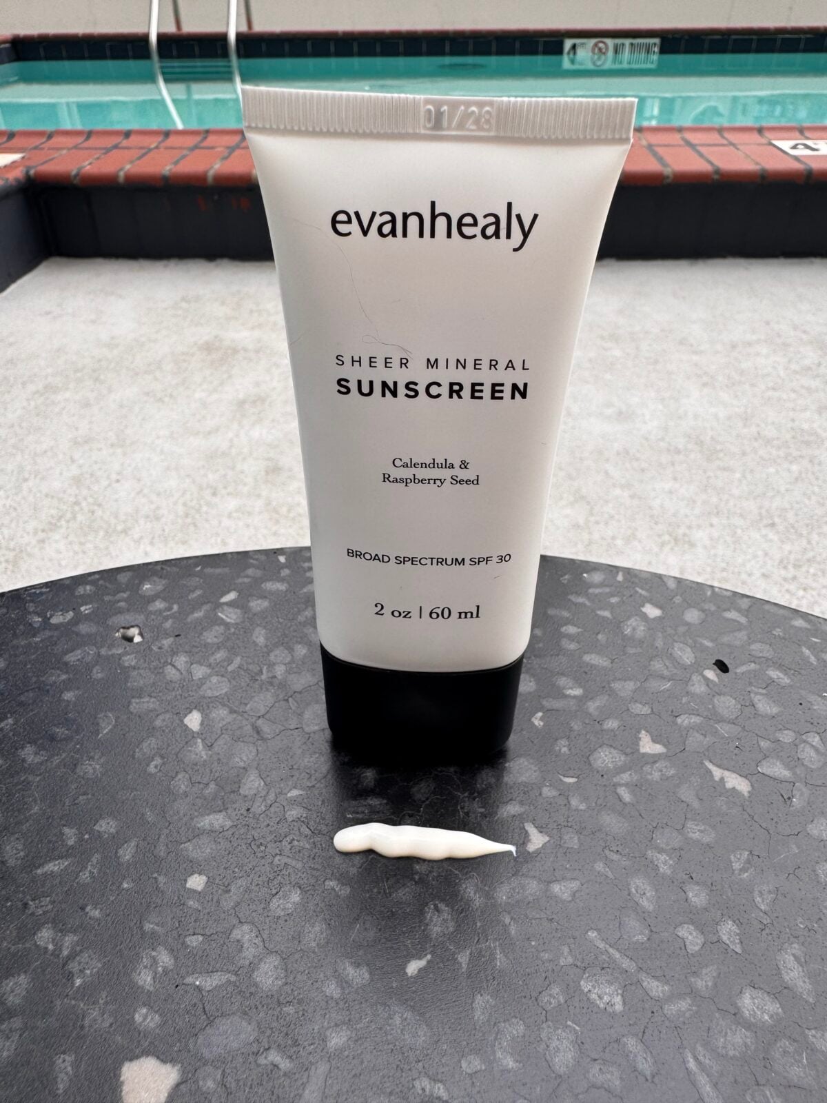 A tube of evanhealy Sheer Mineral Sunscreen stands upright on a black metal table, with a small line of sunscreen in front of it. There is a swimming pool and red brick trim in the background.