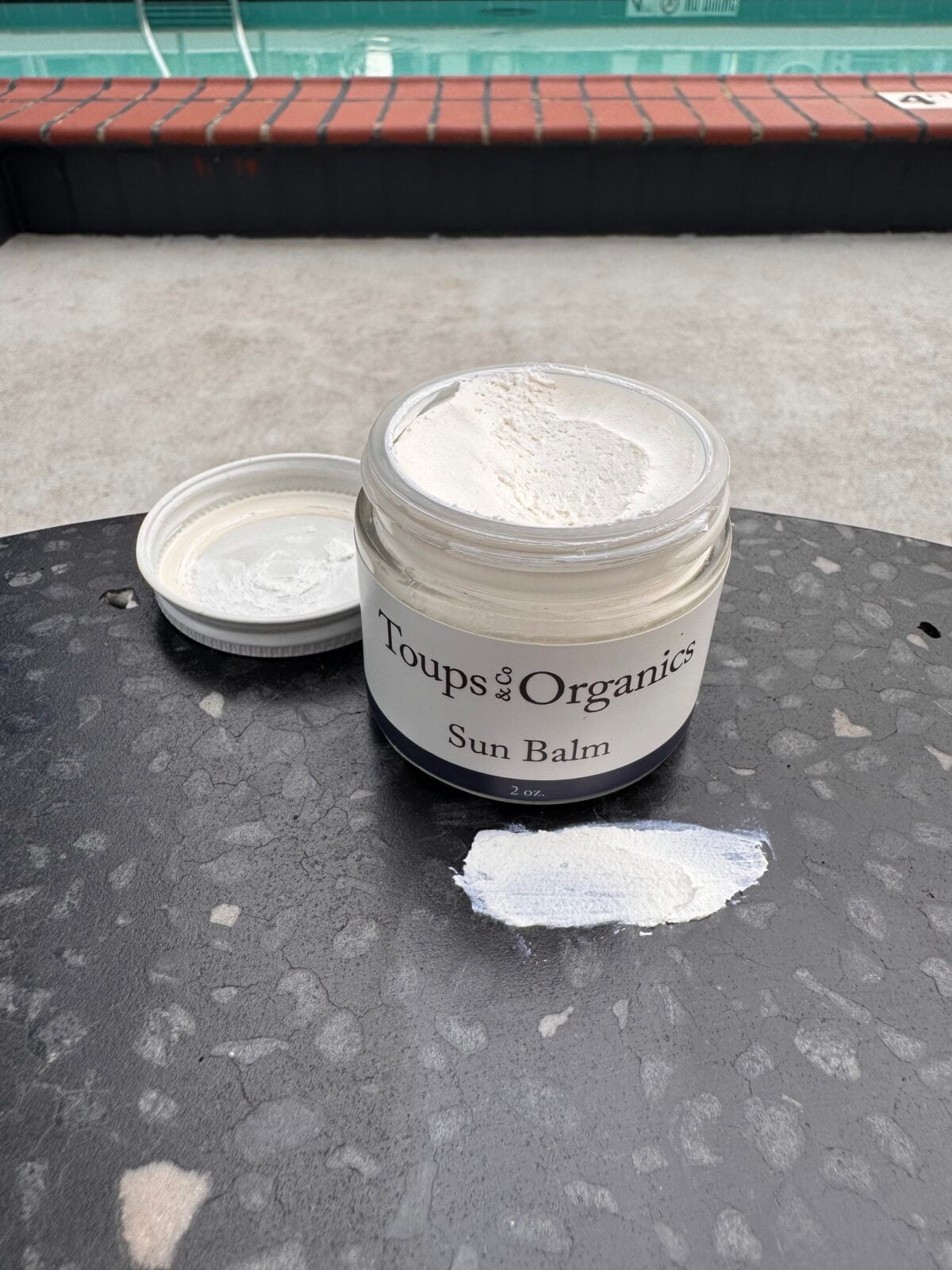 An open jar labeled Toups & Co. Organics Sun Balm sits on a round, dark table near a pool, with its lid off and a small amount of the balm spread on the table surface.