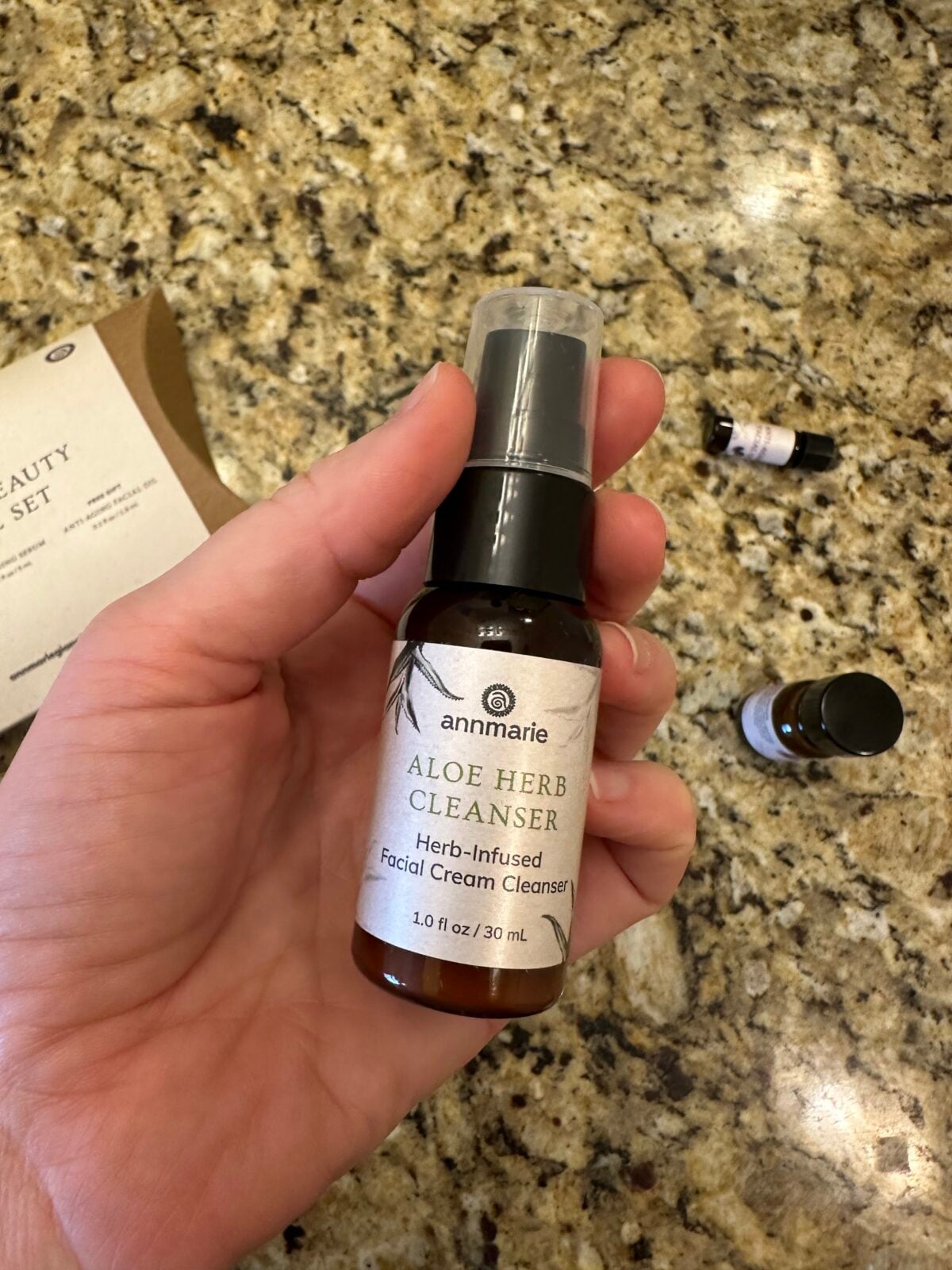 A hand holds a 1 oz bottle of Annmarie Aloe Herb Cleanser on a granite countertop, with another small bottle and packaging visible in the background.