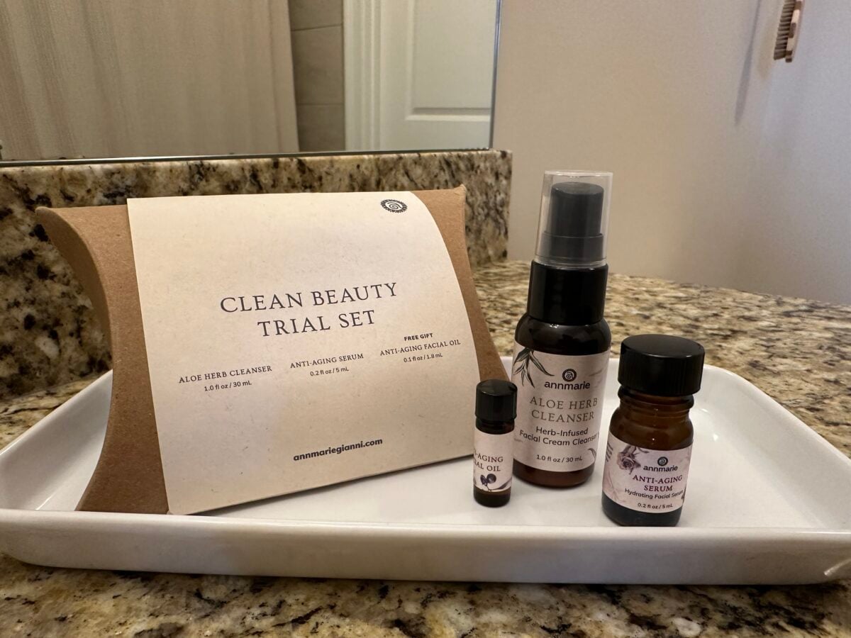 A set of four skincare products labeled Clean Beauty Trial Set is displayed on a white tray atop a granite counter. The set includes cleanser, serum, facial oil, and a small sample vial. A bathroom mirror is in the background.