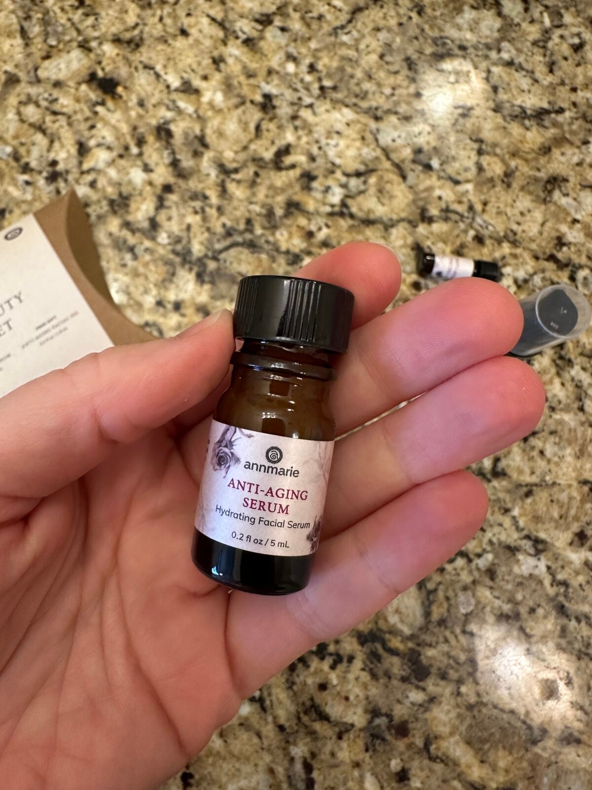 A hand holds a small brown glass bottle labeled annmarie ANTI-AGING SERUM Hydrating Facial Serum 0.2 fl. oz./5 mL over a granite countertop. Another small bottle and packaging are visible in the background.
