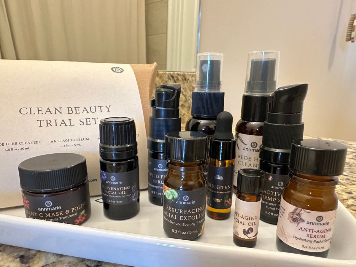 A collection of nine small skincare bottles and jars from Annmarie, labeled as a Clean Beauty Trial Set, displayed on a bathroom counter near a sink with a beige tiled wall in the background.