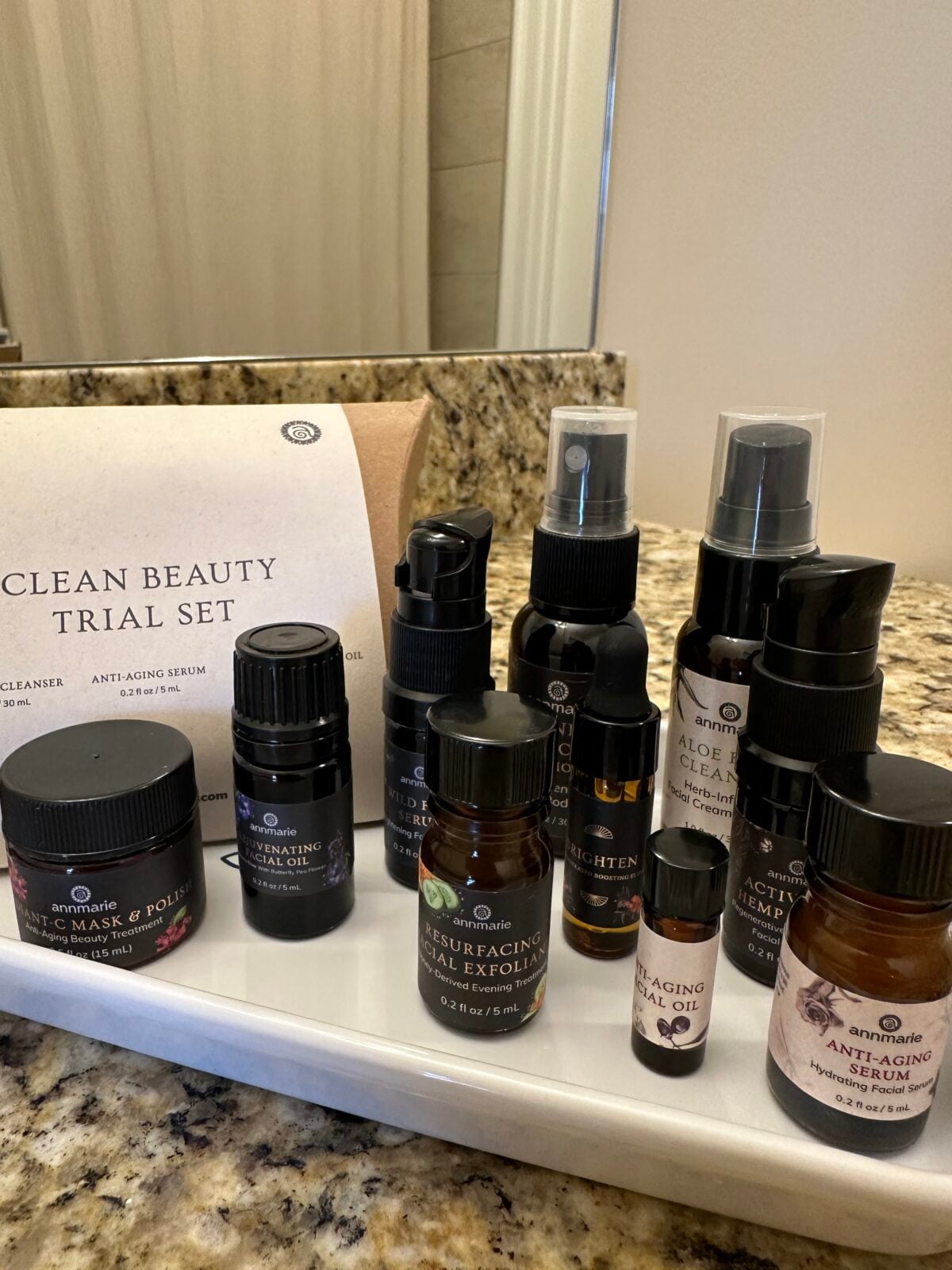 A collection of skincare bottles and containers labeled with various product names is arranged on a white tray on a bathroom counter, next to a card titled Clean Beauty Trial Set..