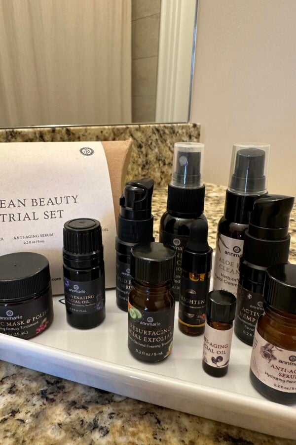 A collection of skincare bottles and containers labeled with various product names is arranged on a white tray on a bathroom counter, next to a card titled Clean Beauty Trial Set..