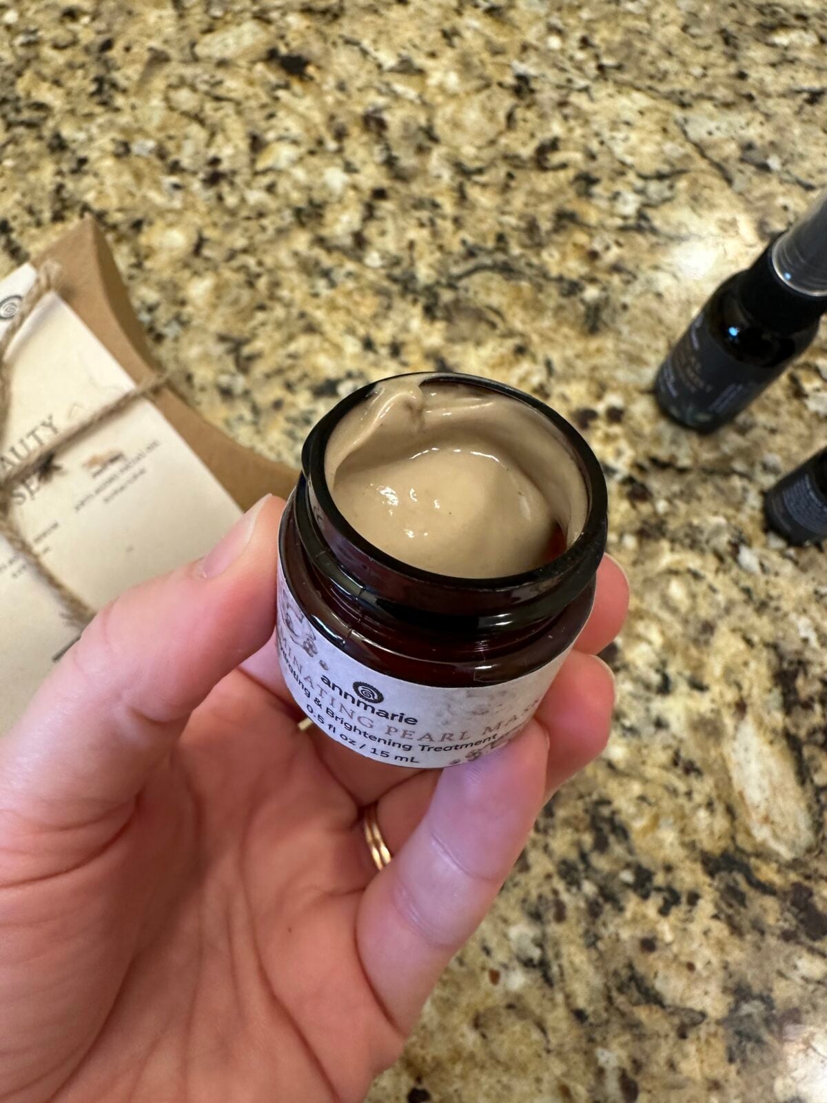 A hand holds an open jar of creamy beige skincare product over a granite countertop, with a cardboard box and two dark bottles visible in the background.