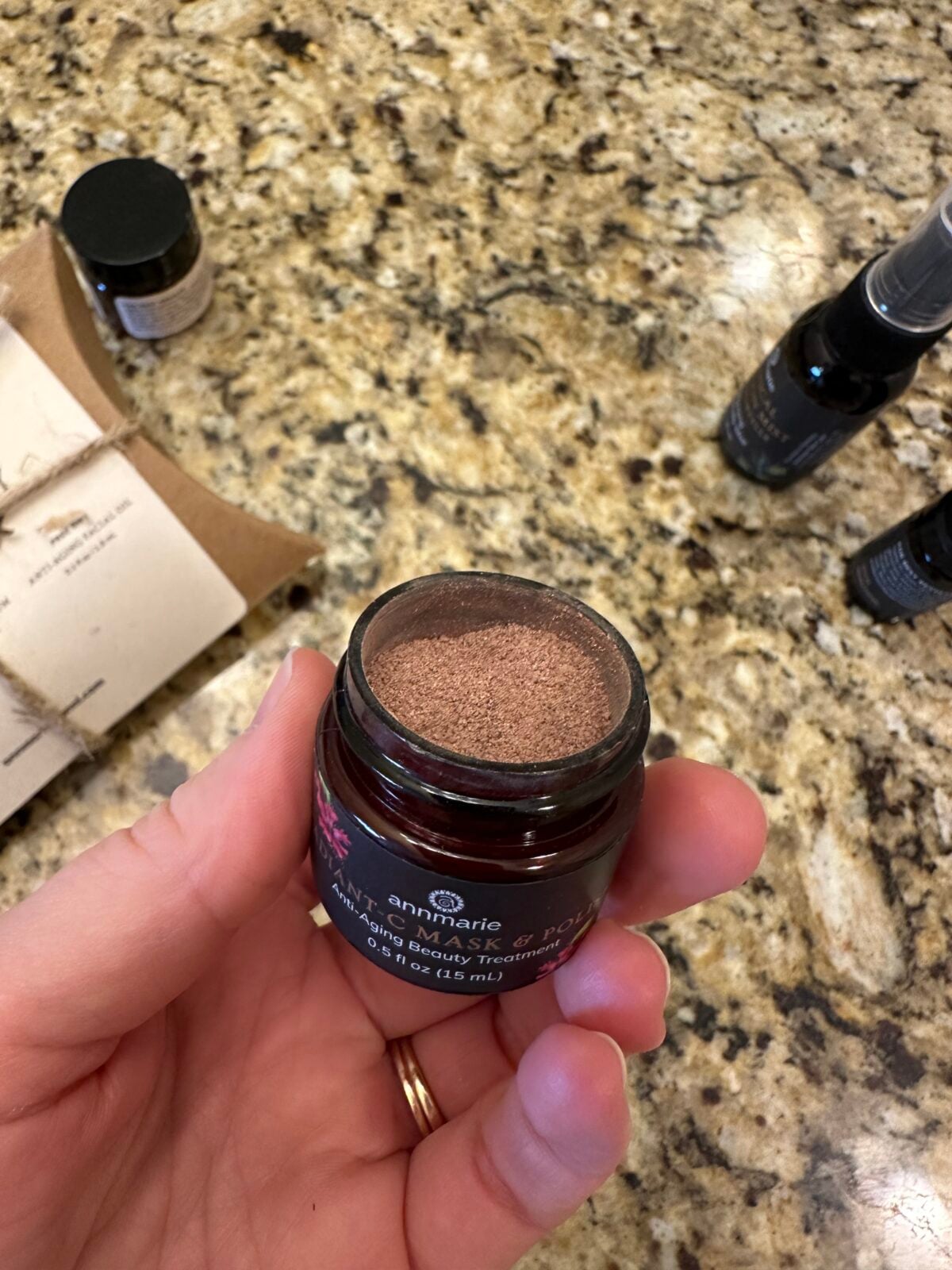 A hand holds an open jar of Annmarie Ayurvedic Mask on a granite countertop, with a product label visible. Other small bottles and a brown paper package are on the counter in the background.