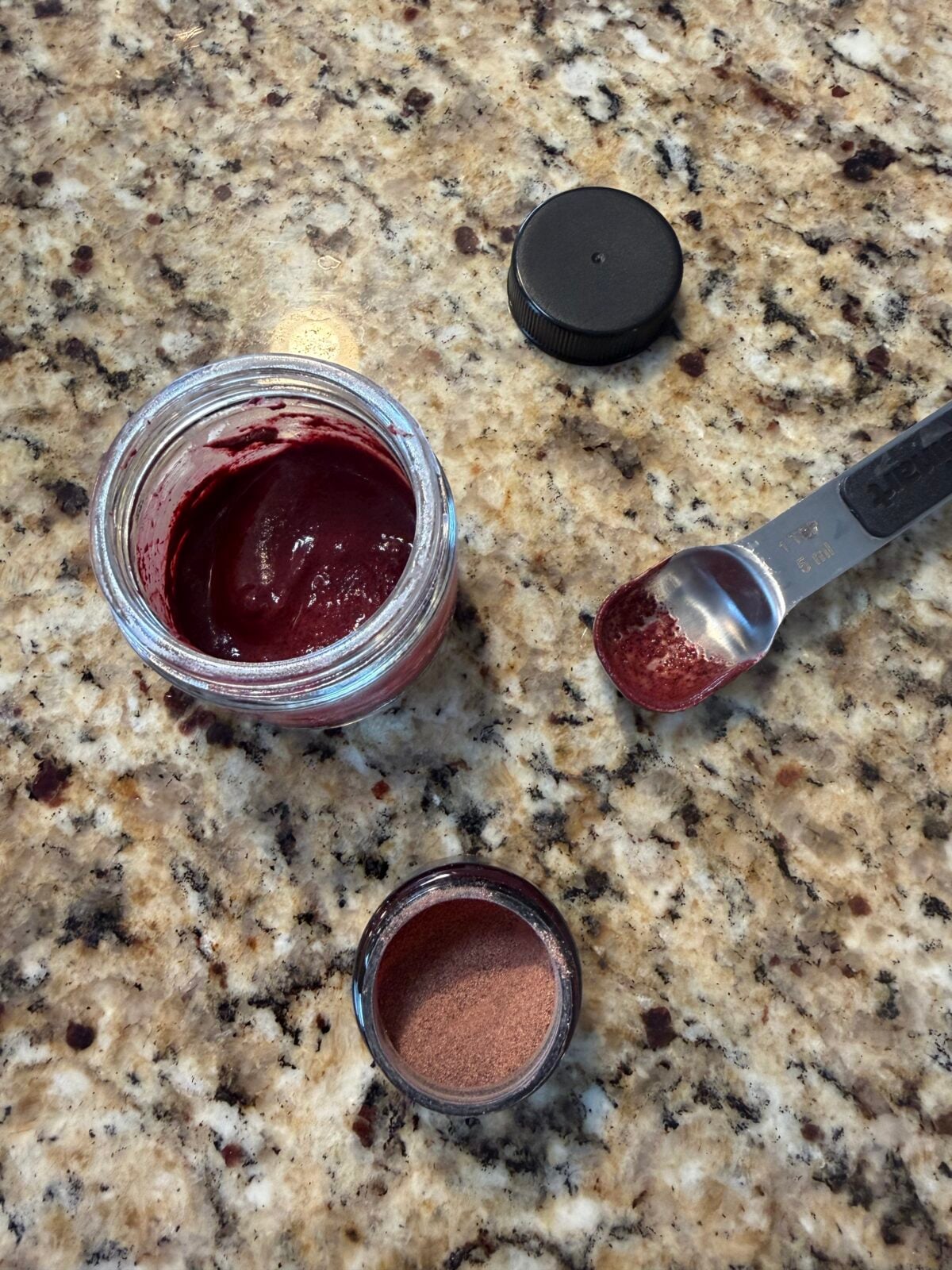 Open jar with dark red paste, a small container with reddish powder, a teaspoon holding more powder, and a black lid are on a speckled granite countertop.