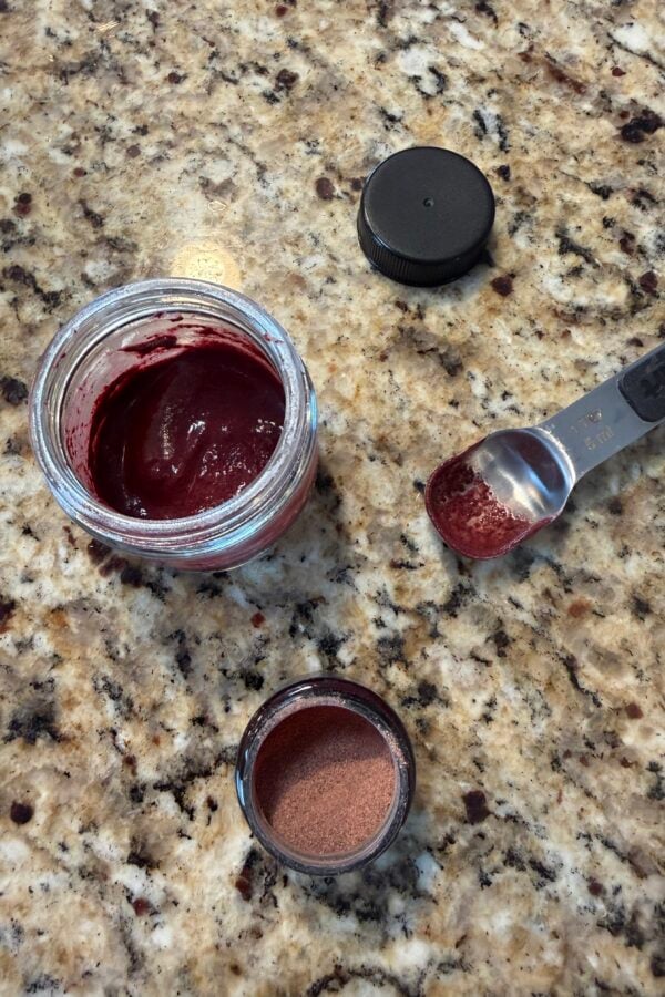 Open jar with dark red paste, teaspoon with powder, small jar with reddish powder, and a black lid on a granite countertop.