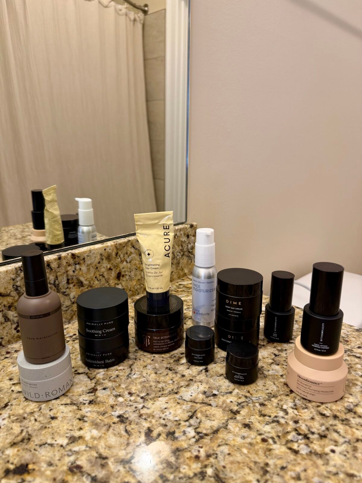 A variety of skincare products, including bottles, jars, and tubes, are neatly arranged on a granite bathroom countertop near a mirror and a beige shower curtain.