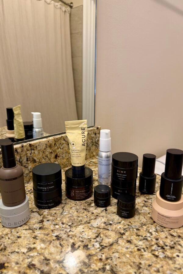 A variety of skincare products, including bottles, jars, and tubes, are neatly arranged on a granite bathroom countertop near a mirror and a beige shower curtain.