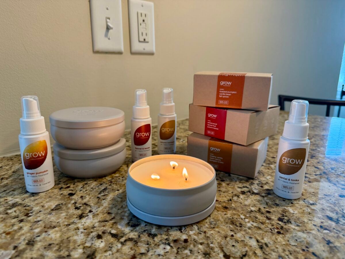 A lit three-wick candle sits on a granite countertop, surrounded by several bottles labeled grow, stacked boxes, and two lidded containers. Electrical outlets and a light switch are visible on the wall in the background.