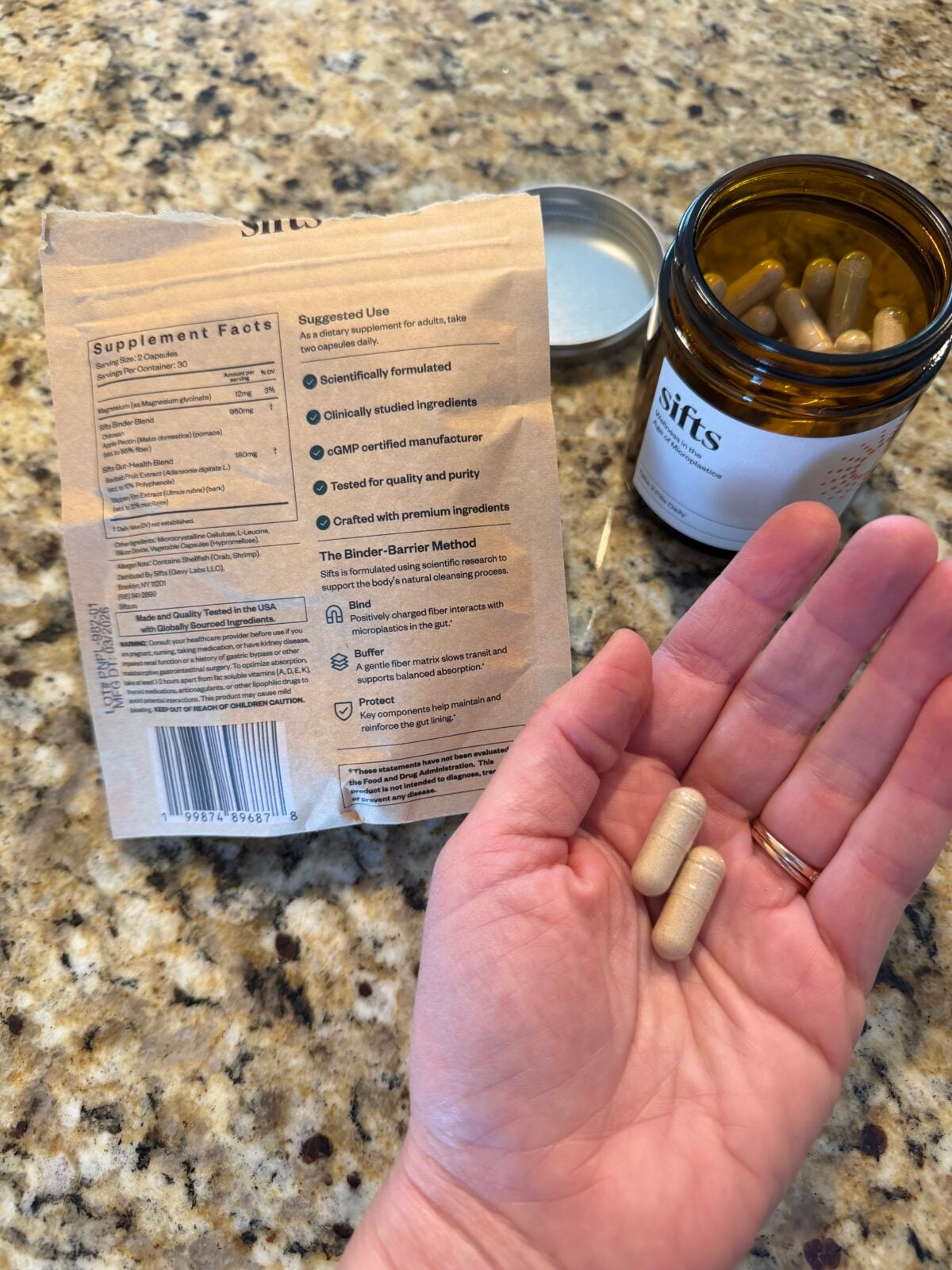 A hand holds two beige capsules above a granite countertop. A brown pill bottle with a metal lid and a packet showing supplement facts are also visible. The bottle is labeled sifts..