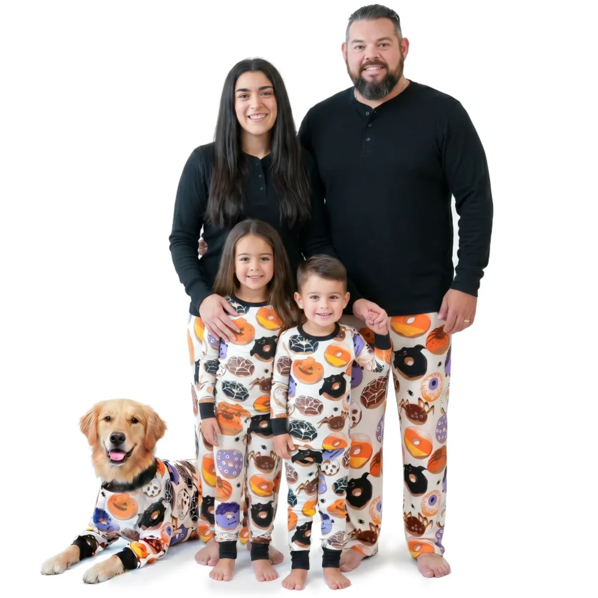 A family of four and a golden retriever dog wear matching Halloween-themed pajamas with pumpkins, bats, and donuts, standing and sitting against a white background.