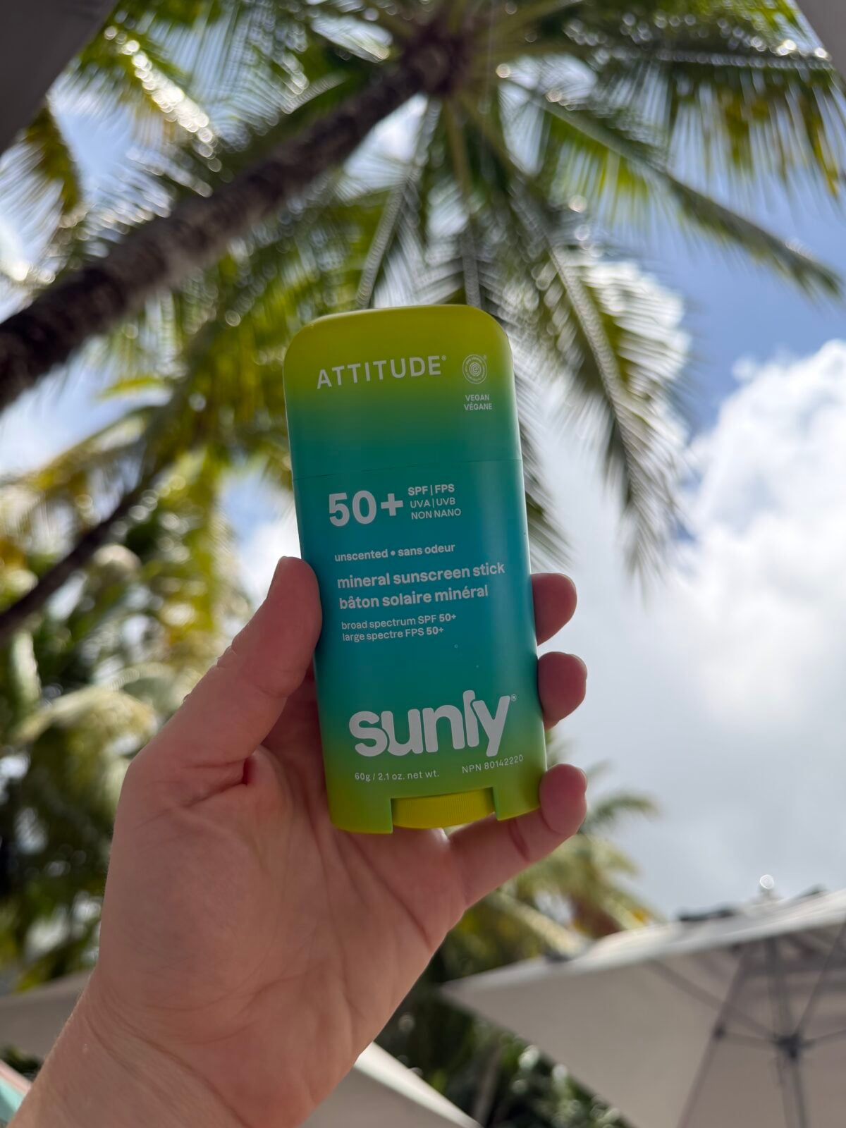 A hand holds a green container of Attitude brand Sunly mineral sunscreen stick SPF 50+ outdoors, with palm trees, blue sky, and some clouds in the background.
