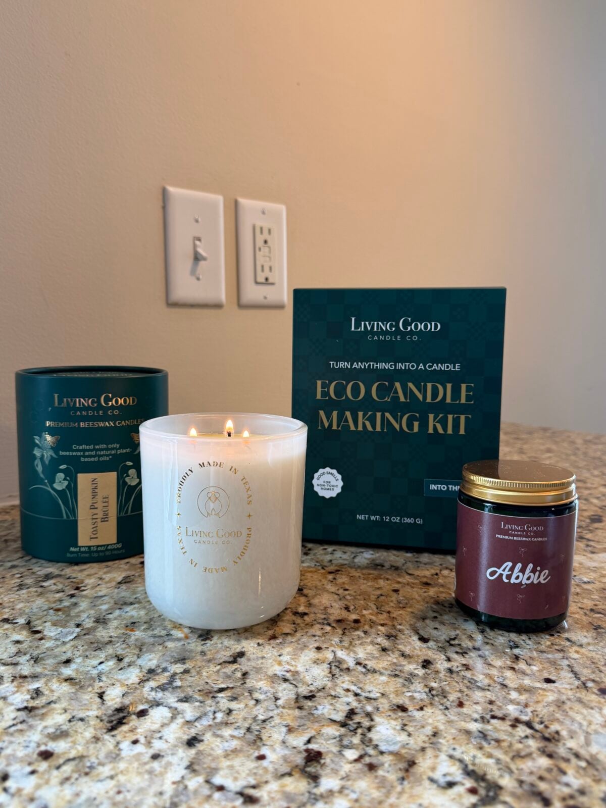 A white candle in a glass jar, a green cylindrical container, an eco candle making kit box, and a small jar labeled Abbie are arranged on a speckled granite countertop near a beige wall with electrical outlets.