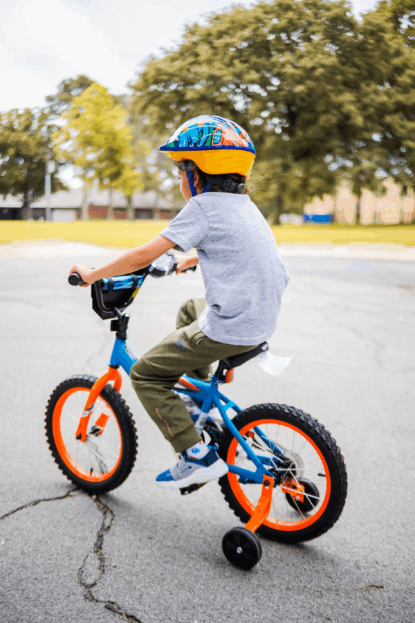 A young child wearing a colorful helmet rides a blue and orange bicycle with training wheels on a paved outdoor surface. Trees and a building are visible in the background.