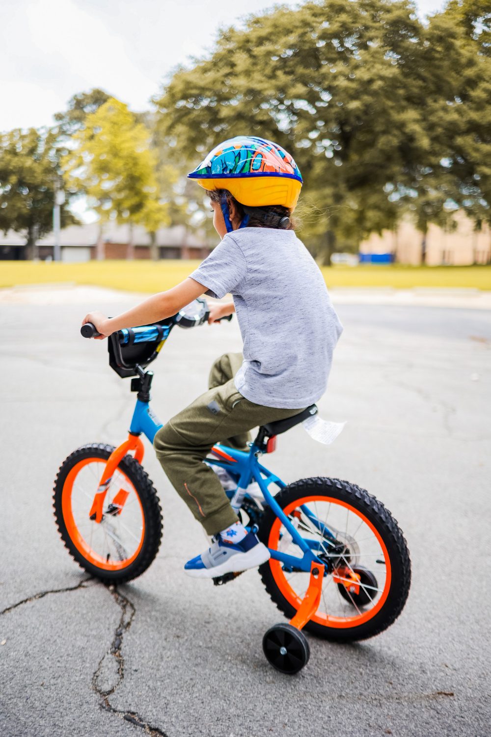 A young child wearing a colorful helmet rides a blue and orange bicycle with training wheels on a paved outdoor surface. Trees and a building are visible in the background.