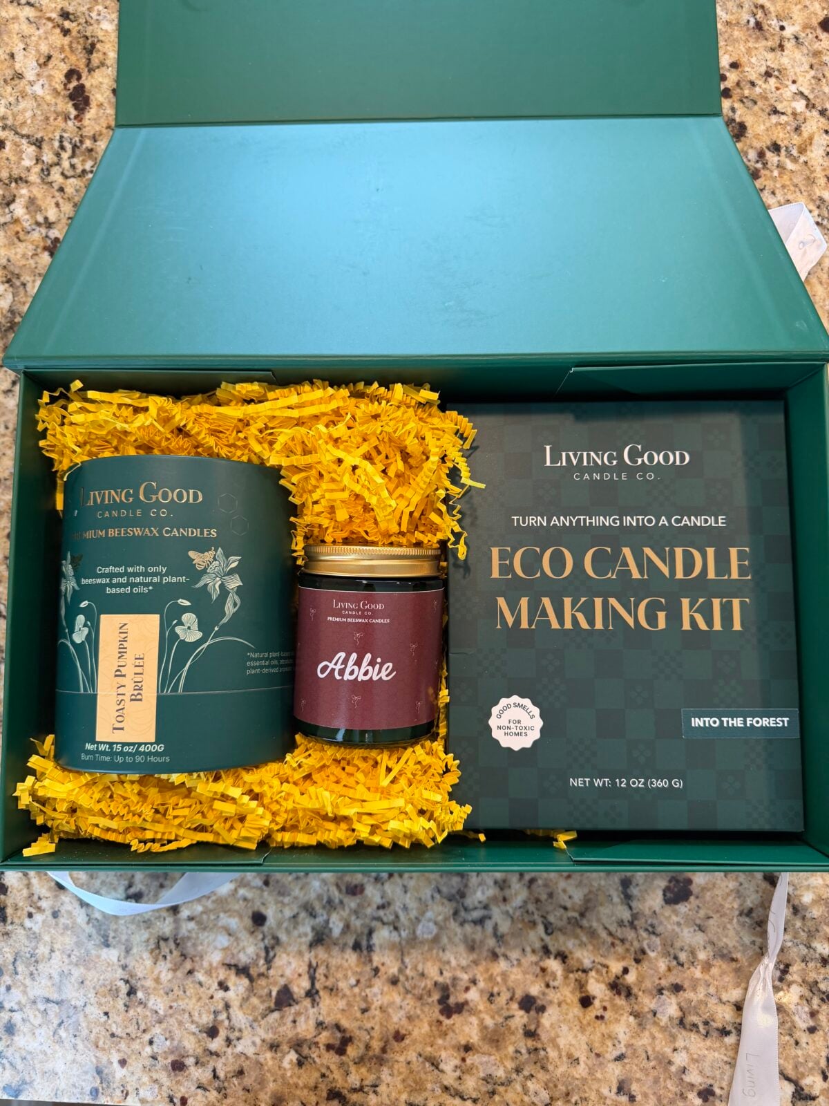 Open green box on granite counter contains yellow shredded paper, a Living Good scented candle, an Abbie labeled small jar, and an Eco Candle Making Kit labeled Into the Forest..