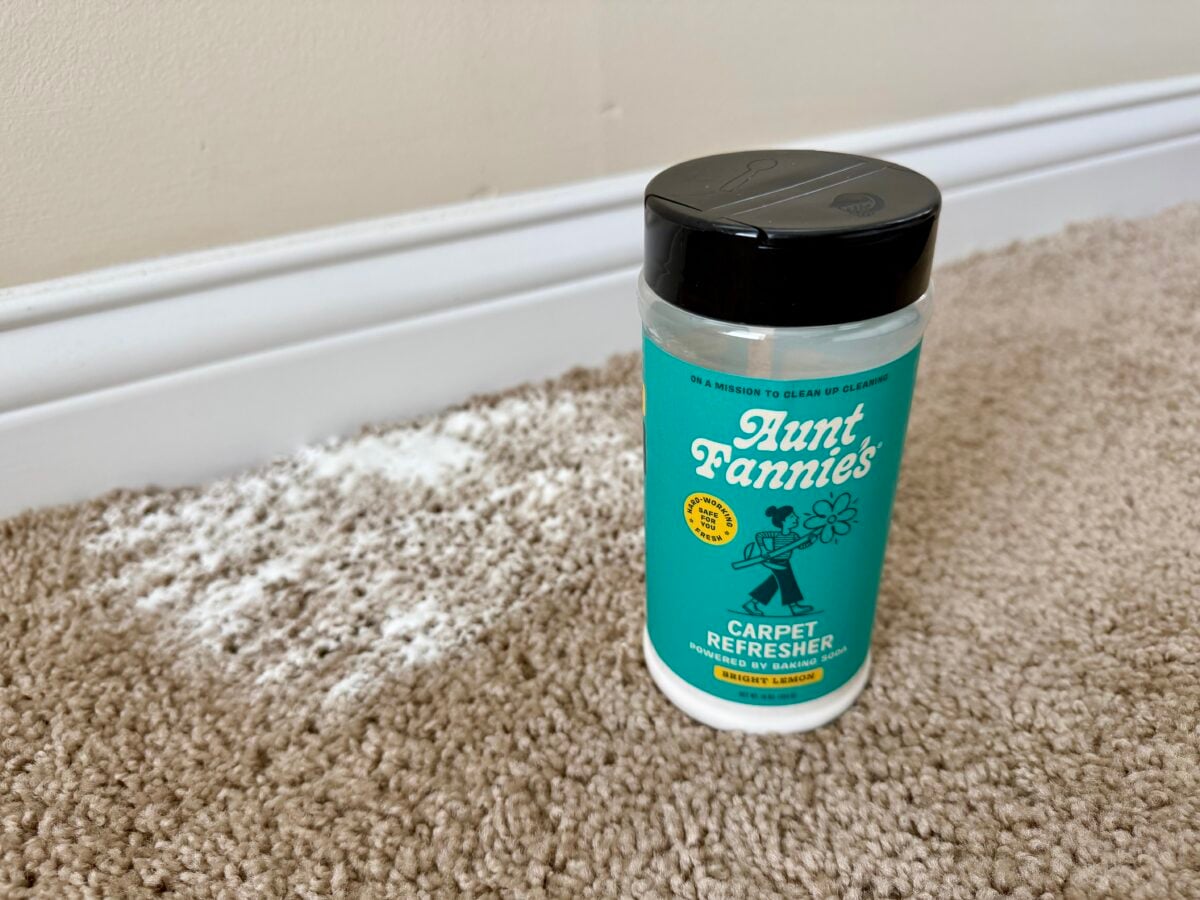 A container of Aunt Fannies Carpet Refresher sits on beige carpet with white powder sprinkled nearby. The wall and white baseboard are visible in the background.