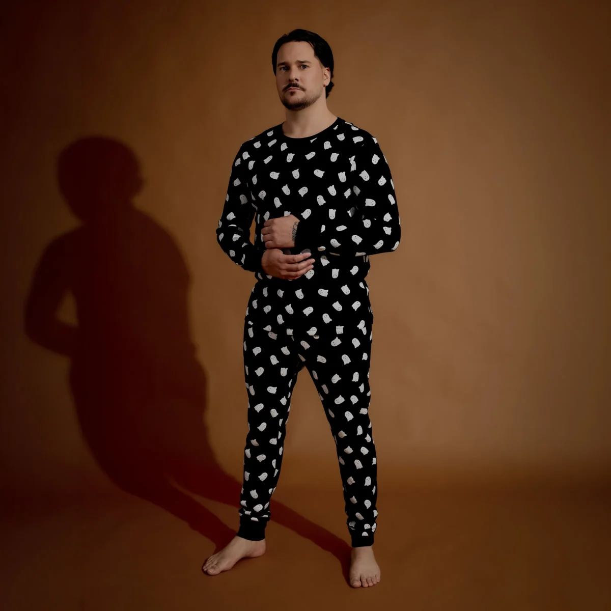 A person stands barefoot on a brown floor with a brown background, wearing a black long-sleeve pajama set with a white abstract pattern, looking forward with hands clasped at the waist.